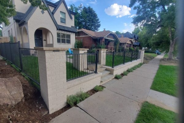 Steel wrought iron fence Denver - A Straight Up Fence Company Residential street scene: beige brick pillars with black metal railing along a front-yard fence, sidewalk in foreground and houses beyond.